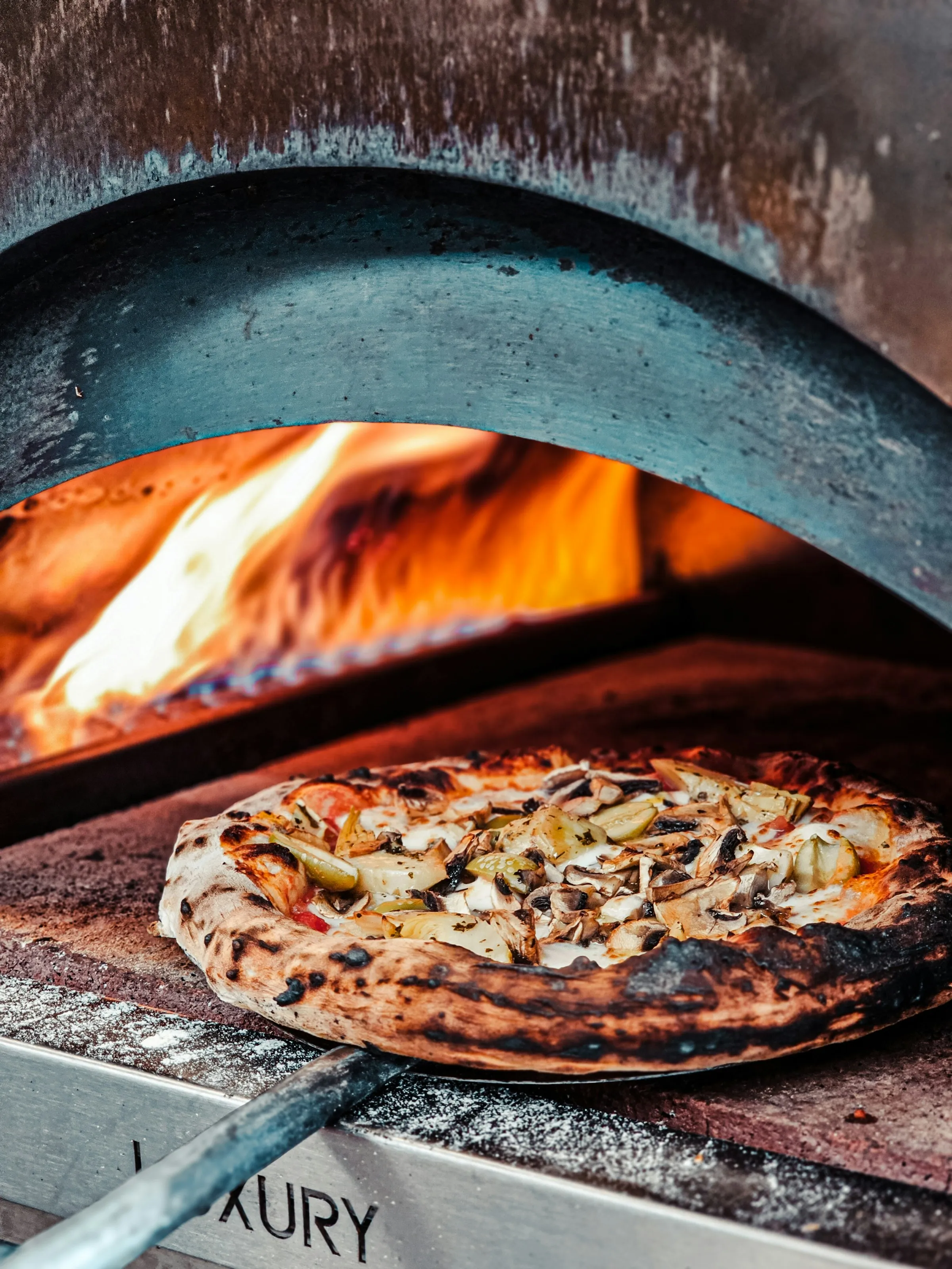A charred pizza on a peel at the mouth of a lit wood-fired oven.